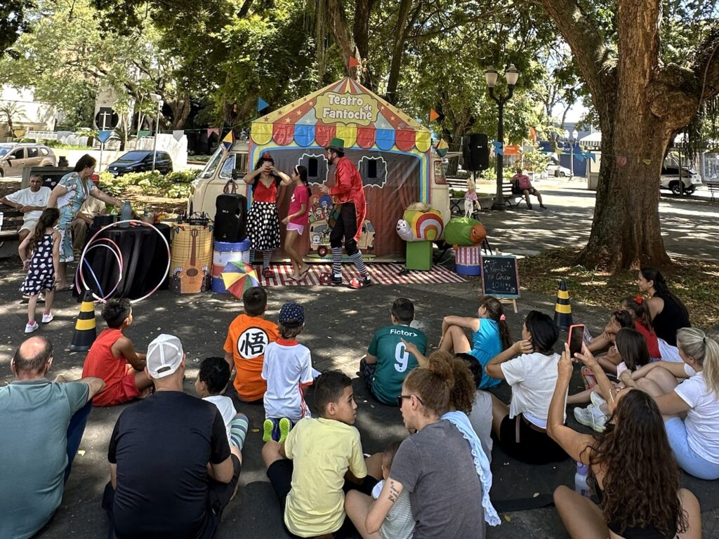 A cena retrata uma apresentação de teatro de fantoches ao ar livre, provavelmente em um parque ou praça, sob a sombra de árvores frondosas. No centro da imagem, um pequeno palco improvisado, decorado com cores vibrantes e a inscrição "Teatro de Fantoche", serve de cenário para a performance. Um homem vestido de palhaço, com roupas vermelhas e chapéu verde, interage com duas crianças que parecem ser as atrizes. Uma mulher com uma blusa estampada e uma menina com vestido xadrez observam a cena atentamente, enquanto um homem mais velho, sentado em um banco ao fundo, também parece acompanhar o espetáculo. À frente do palco, um público composto majoritariamente por crianças e alguns adultos está sentado no chão, assistindo à apresentação. As crianças vestem roupas casuais e coloridas, algumas com estampas de personagens infantis. Uma mulher na plateia está registrando o momento com seu celular. Ao redor do palco, há elementos que remetem a um circo ou festival, como Hula Hoops, um guarda-chuva colorido e caixas decoradas com instrumentos musicais. Ao fundo, é possível avistar carros estacionados e edifícios, indicando que a apresentação ocorre em uma área urbana. A luz do sol, filtrada pelas árvores, cria um ambiente agradável e descontraído.