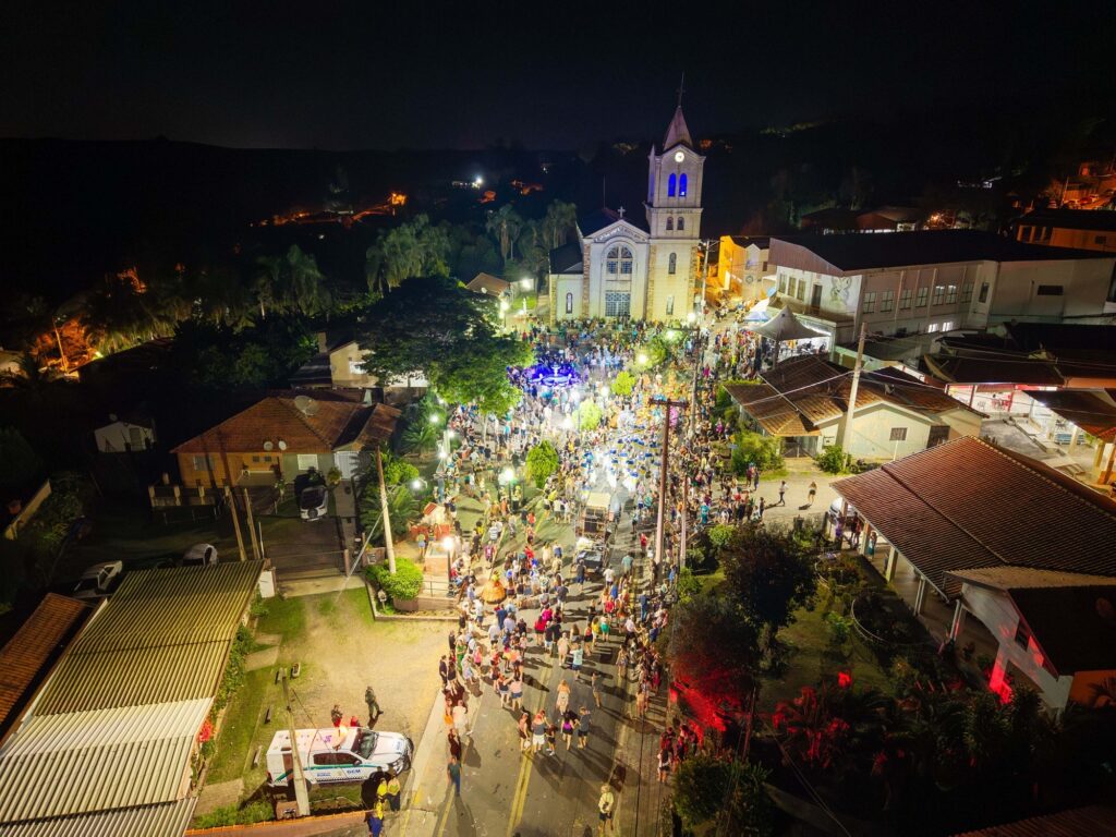 A imagem é uma vista aérea noturna de uma rua movimentada, iluminada por postes de luz e pelas luzes das casas e estabelecimentos comerciais. No centro da cena, destaca-se uma igreja com uma torre alta, cujos sinos estão iluminados em azul. A rua está repleta de pessoas, que parecem estar participando de uma festa ou evento, com algumas pessoas vestidas com fantasias coloridas. Há também carros e barracas montadas ao longo da rua, sugerindo uma feira ou celebração. As casas ao redor têm telhados de barro e algumas janelas iluminadas, criando um ambiente acolhedor. Ao fundo, a paisagem é escura, com algumas luzes distantes indicando a presença de outras construções. A atmosfera geral é de celebração e comunidade.