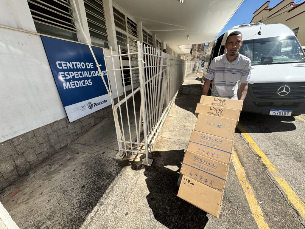 A imagem mostra um homem de pé em frente a uma van branca, carregando uma pilha de caixas de papelão. Ele está vestindo uma camisa polo cinza com listras azuis e vermelhas. As caixas estão empilhadas em uma escada e parecem ser entregas médicas, com os logotipos "biolab & CO." e "CRISTÁLIA" visíveis. Atrás do homem, há um prédio com uma placa azul que diz "CENTRO DE ESPECIALIDADES MÉDICAS" e "SMS SECRETARIA DE SAÚDE Piracicaba". A cena parece ser em um dia ensolarado, com sombras projetadas no chão. A van tem uma placa de licença brasileira visível.