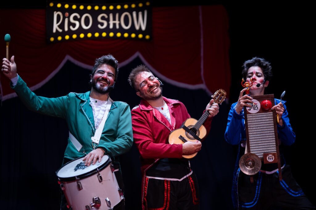 A imagem mostra três artistas de circo no palco, com uma placa escrita "RISO SHOW" ao fundo. Os artistas estão vestidos com roupas coloridas e maquiagem de palhaço. O artista da esquerda, vestindo um casaco verde, está segurando uma baqueta e tocando um tambor. Ele tem uma barba e um sorriso largo no rosto. O artista do meio, com um casaco vermelho, está tocando um pequeno violão. Ele também está sorrindo e parece estar cantando ou rindo. A artista da direita, vestindo um casaco azul, está tocando uma tábua de lavar como instrumento musical, e tem um pequeno boneco de macaco em uma das mãos. Ela também está maquiada e parece estar engajada na performance. O fundo do palco é escuro, com cortinas vermelhas e uma luz amarelada vinda da placa "RISO SHOW". A atmosfera geral da imagem sugere uma apresentação animada e divertida.