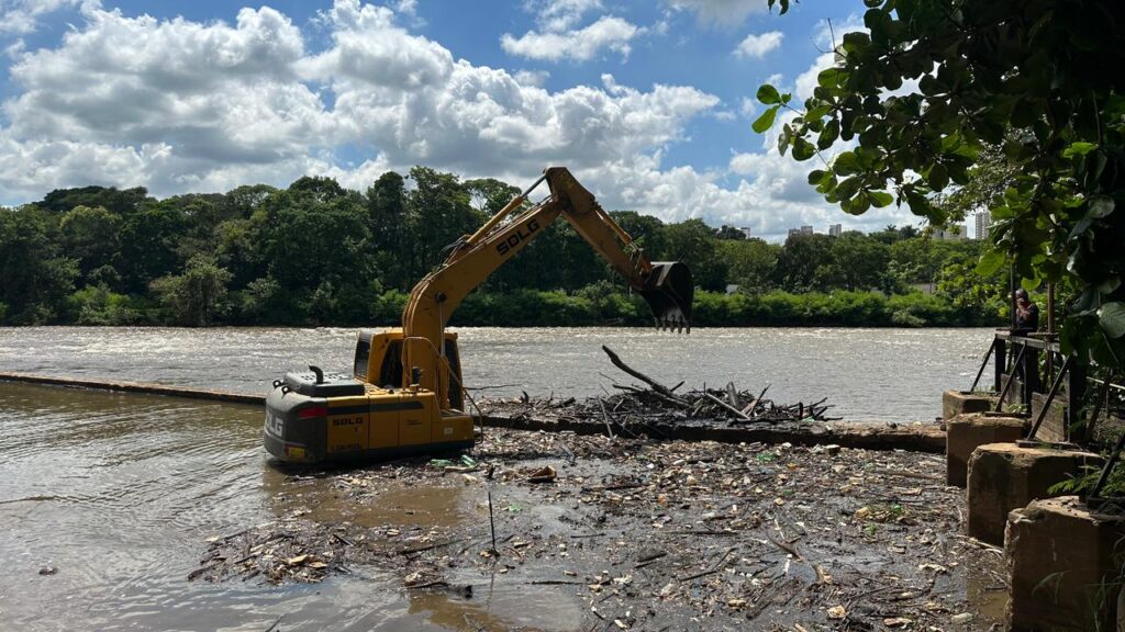 A imagem mostra um escavador amarelo da marca SDLG posicionado à beira de um rio, utilizando sua concha para retirar galhos e resíduos acumulados na água. O fluxo do rio é contínuo e há uma barreira instalada para conter ou direcionar os detritos. Do outro lado da margem, vê-se uma área verde com árvores densas e, ao fundo, alguns prédios urbanos. À direita, uma pessoa observa a operação a partir de uma plataforma. O céu parcialmente nublado, com aberturas de azul, reforça o clima de dia ensolarado em uma ação de limpeza e manutenção ambiental.