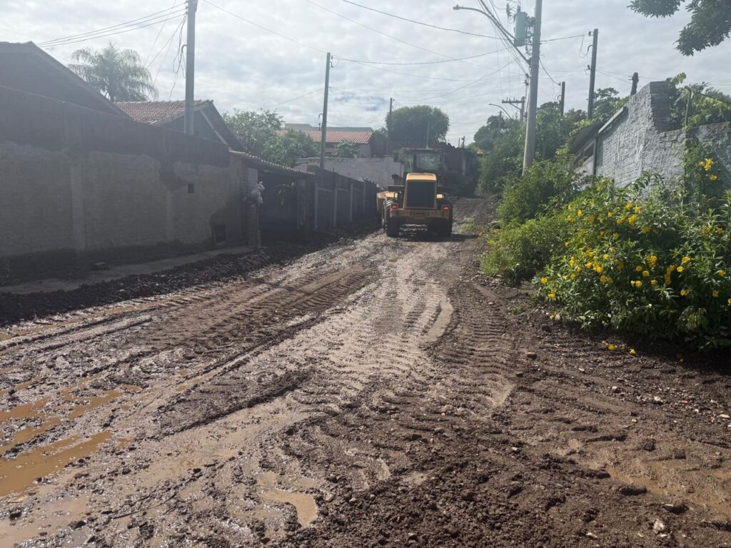 A imagem mostra uma rua de terra muito lamacenta, com marcas profundas de pneus de um trator ou máquina similar. A lama parece ter sido revolvida recentemente, com montes de terra espalhados. À esquerda, vemos casas com muros altos e telhados aparentes, algumas com aspecto mais antigo e outras mais novas. A vegetação é escassa nas proximidades imediatas das casas, mas há algumas árvores e arbustos ao fundo. À direita, a vegetação é mais abundante, com um arbusto carregado de flores amarelas vibrantes que se destaca em meio à terra e à lama. Uma construção em ruínas ou em fase de construção, com paredes de tijolos aparentes, também está presente à direita. Ao fundo, a rua continua subindo, e podemos ver a traseira de uma máquina amarela, provavelmente um trator ou retroescavadeira, que parece estar trabalhando no local. Fios elétricos e postes de luz atravessam o céu nublado, indicando que se trata de uma área urbana ou suburbana.