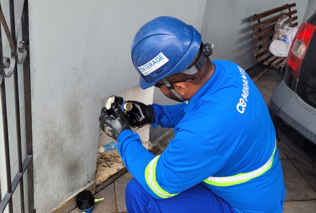 Um trabalhador vestindo um uniforme azul, capacete azul e luvas pretas está agachado, examinando um hidrômetro. O uniforme tem listras refletivas amarelas e brancas nos braços e na cintura. No capacete, há um adesivo branco com texto azul que diz "INTERAGE". No lado direito da camisa, em letras brancas estilizadas, está escrito "MIRANTE". O trabalhador está com as mãos perto de uma tubulação de água azul que sai de uma parede cinza. Ao fundo, há um banco de madeira com alguns objetos, incluindo sacolas plásticas e um rolo de tecido branco. Um carro escuro está estacionado ao lado.