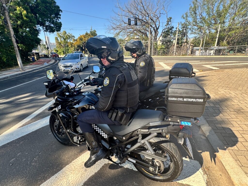 A imagem mostra dois guardas civis metropolitanos em suas motocicletas, parados em uma rua. Ambos estão vestindo uniformes pretos, coletes táticos e capacetes. As motocicletas são pretas e equipadas com caixas de armazenamento traseiras que exibem a inscrição "GUARDA CIVIL METROPOLITANA". Um dos guardas está mais próximo da câmera, com o rosto parcialmente visível sob o capacete. O outro guarda está um pouco mais atrás, também em sua motocicleta. O cenário parece ser urbano, com carros passando na rua e árvores ao fundo. O céu está azul e ensolarado, indicando um dia claro.