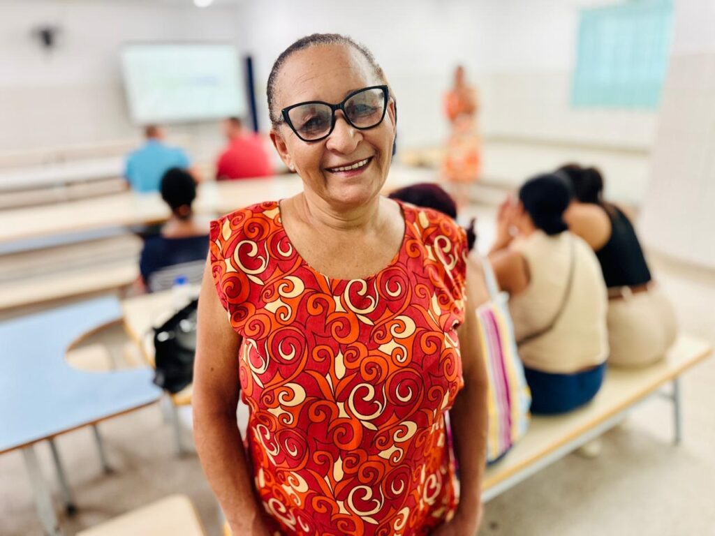 A imagem mostra uma mulher mais velha sorrindo para a câmera, usando óculos pretos e uma blusa vermelha vibrante com um padrão laranja e amarelo. Ela está em primeiro plano, com o fundo desfocado, onde se veem outras pessoas sentadas em mesas e bancos em uma sala que parece ser uma sala de aula ou auditório. Há um quadro branco ao fundo, com algumas anotações. A iluminação é clara e parece ser luz natural.