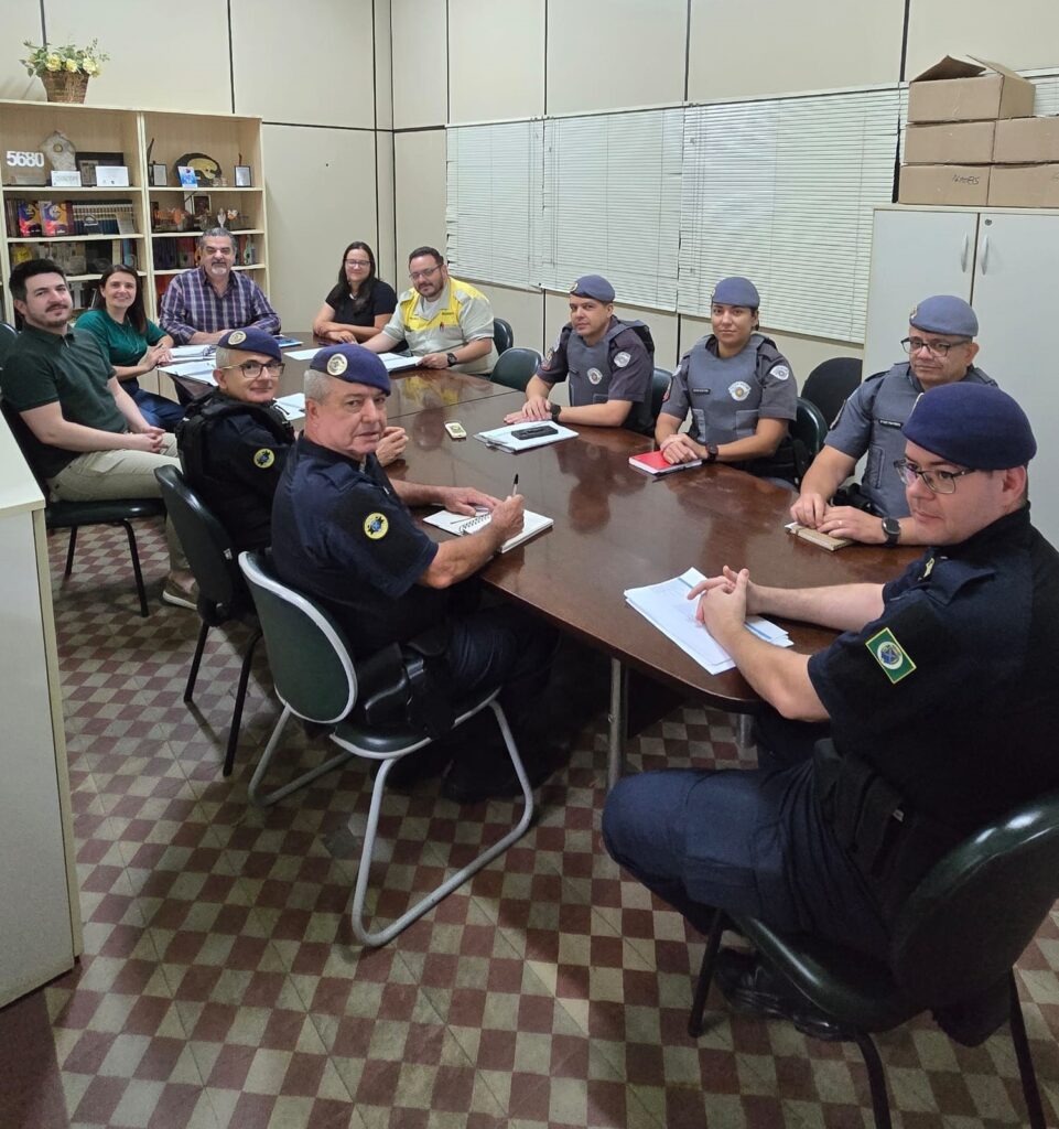 O grupo de pessoas está reunido em uma sala de reuniões, sentados ao redor de uma mesa de madeira. Há um total de dez pessoas na imagem, a maioria delas vestindo uniformes azuis escuros com boinas correspondentes, sugerindo que são membros de uma força policial ou militar. Alguns estão olhando para a frente, enquanto outros estão engajados em conversas ou tomando notas. Há papéis e cadernos sobre a mesa, indicando que uma reunião ou sessão de treinamento está em andamento. O ambiente parece ser um escritório ou sala de conferências, com estantes cheias de livros e objetos decorativos em uma parede e persianas em outra. O piso tem um padrão xadrez distinto em tons de marrom e bege.