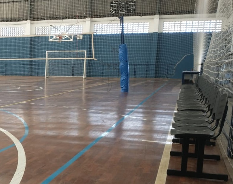A imagem mostra o interior de uma quadra poliesportiva. No centro, há uma quadra de madeira com marcações de linhas azuis e brancas, indicando as áreas de jogo para diferentes esportes. Uma rede de vôlei está estendida horizontalmente, dividindo a quadra ao meio. Ao fundo, é possível ver uma tabela de basquete e um gol, sugerindo que a quadra é utilizada para diversas modalidades esportivas. As paredes laterais são cobertas por uma tela de proteção azul. Na lateral direita da imagem, uma fileira de assentos pretos está disposta ao longo da quadra. O teto da quadra é alto, com iluminação embutida e uma estrutura metálica visível.