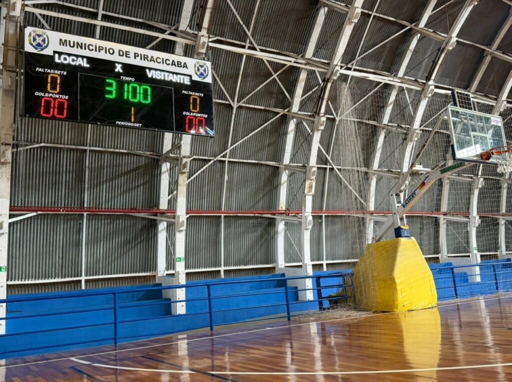 A imagem mostra o interior de uma quadra de esportes, provavelmente de basquete, com foco em um placar eletrônico. O placar exibe informações do jogo, incluindo o nome do município "MUNICÍPIO DE PIRACICABA", as pontuações das equipes "LOCAL" e "VISITANTE", o tempo de jogo e o período. No placar, a equipe "LOCAL" tem 0 faltas/sets e 00 gols/pontos. A equipe "VISITANTE" também tem 0 faltas/sets e 00 gols/pontos. O tempo de jogo está marcado como 31:00, e o período/set atual é 1. A quadra possui piso de madeira polida, com marcações brancas. Ao fundo, há arquibancadas azuis com grades e uma estrutura metálica que sustenta o teto e a iluminação. Um aro de basquete com sua tabela de vidro está visível à direita, com uma proteção amarela acolchoada na base do suporte. A iluminação parece ser artificial, vinda do teto.