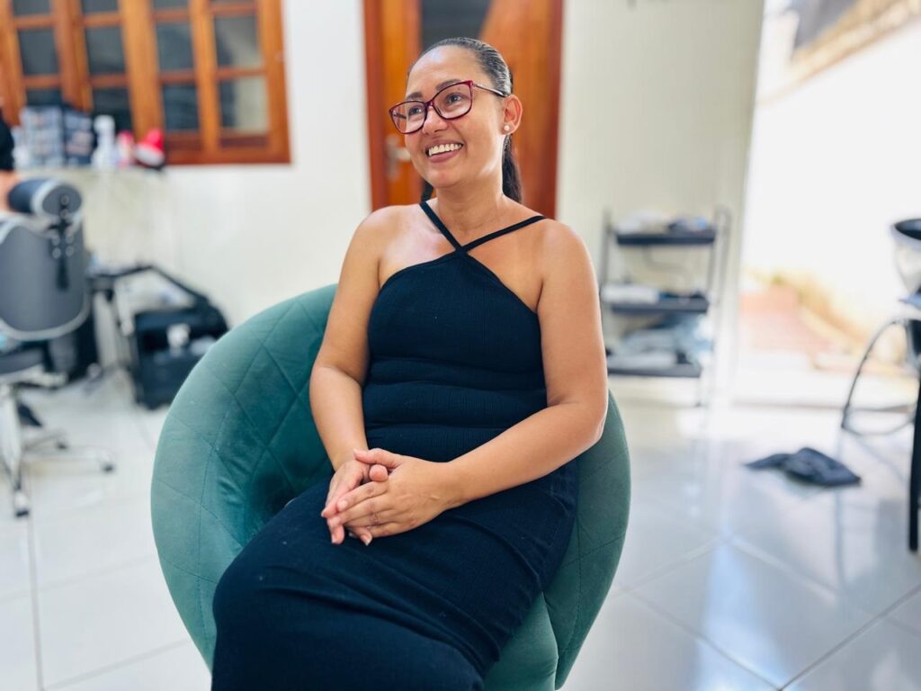 A mulher está sentada em uma cadeira azul-petróleo, vestindo um vestido preto justo com alças finas. Ela usa óculos de aro vermelho e tem o cabelo preso em um rabo de cavalo. As mãos dela estão entrelaçadas no colo. O fundo da imagem está desfocado, mas é possível ver uma janela com moldura de madeira e prateleiras com objetos.