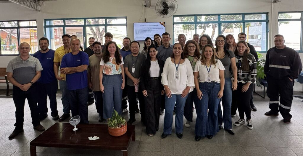 A imagem mostra um grupo de aproximadamente 25 pessoas reunidas em um ambiente interno, iluminado por luz natural que entra pelas janelas ao fundo. O grupo está posicionado em três fileiras, com pessoas em pé e algumas um pouco à frente. A maioria está sorrindo e olhando para a câmera, transmitindo um clima amistoso e de confraternização. A diversidade do grupo é notável, com homens e mulheres de diferentes idades e estilos de roupas, que variam entre uniformes azuis com o logo "Semae" e roupas casuais ou formais, como blusas brancas e um blazer preto. O local parece ser um escritório ou uma instituição, com piso de cerâmica clara, paredes brancas e uma mesa baixa de madeira na frente do grupo, onde há um vaso com uma planta, um recipiente de vidro e alguns papéis. Na parede ao fundo, há uma tela digital exibindo uma lista intitulada "REFERÊNCIAS BIBLIOGRÁFICAS", contendo citações de obras relacionadas à saúde mental e políticas públicas, indicando que o grupo pode estar envolvido em atividades acadêmicas, profissionais ou de formação na área de saúde mental ou assistência social. No geral, a foto transmite uma imagem de equipe, colaboração e profissionalismo em um ambiente acolhedor e organizado.
