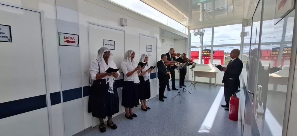 A imagem mostra um grupo de pessoas em um corredor branco com portas. No lado esquerdo, três mulheres estão em pé, vestindo véus brancos e segurando livros, parecendo cantar ou ler em voz alta. Elas estão alinhadas com as portas ao fundo, uma das quais tem uma placa indicando "WC FEMININO". No lado direito do corredor, um grupo de quatro homens está tocando instrumentos de corda, provavelmente violinos, em pé. Eles estão vestidos com ternos escuros. Um homem em pé à direita, também de terno escuro, segura um livro e parece estar dirigindo ou conduzindo a música. Há um extintor de incêndio vermelho no chão perto dele. O corredor tem uma iluminação clara e janelas grandes que permitem a entrada de luz natural e mostram o exterior, onde se pode ver edifícios com letreiros vermelhos. O ambiente parece ser interno, possivelmente em uma instalação temporária ou modular, dada a construção das paredes.