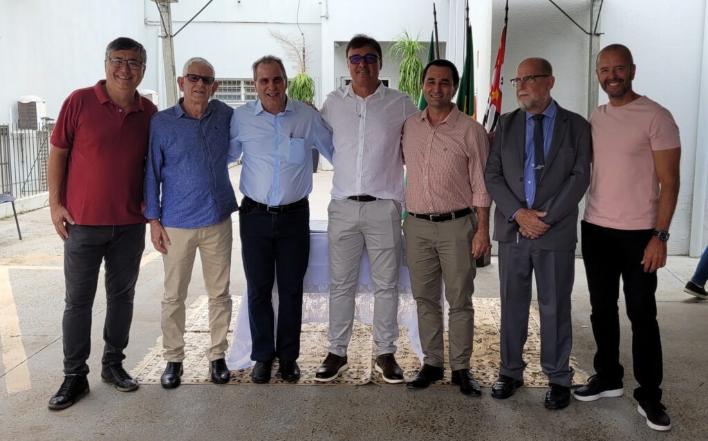 A imagem mostra um grupo de sete homens em pé, alinhados lado a lado, em frente a uma parede branca e algumas bandeiras ao fundo. Eles parecem estar em um evento formal ou reunião. Da esquerda para a direita: 1. O primeiro homem veste uma camisa polo vermelha, calça cinza escura e tênis pretos. Ele usa óculos e tem um sorriso no rosto. 2. O segundo homem está com uma camisa azul de botões, calça bege e sapatos pretos. Ele também usa óculos. 3. O terceiro homem veste uma camisa azul clara de botões, calça azul escura e sapatos pretos. Ele tem um sorriso largo. 4. O quarto homem usa uma camisa branca de botões, calça cinza clara e sapatos marrons. Ele está com os braços levemente levantados, quase abraçando os homens ao lado. 5. O quinto homem veste uma camisa xadrez em tons de rosa e branco, calça bege e sapatos pretos. Ele também tem um sorriso. 6. O sexto homem está de terno cinza escuro, camisa azul clara e gravata azul escura. Ele usa óculos e tem as mãos cruzadas. 7. O sétimo homem veste uma camiseta rosa, calça preta e tênis pretos com detalhes brancos. Ele tem um relógio no pulso esquerdo e um sorriso aberto. Eles estão em uma área externa, com um piso de concreto e um tapete estampado no chão sob os pés. Há uma mesa coberta com uma toalha branca atrás do quarto e quinto homens. As bandeiras ao fundo sugerem um local oficial ou de importância cívica.
