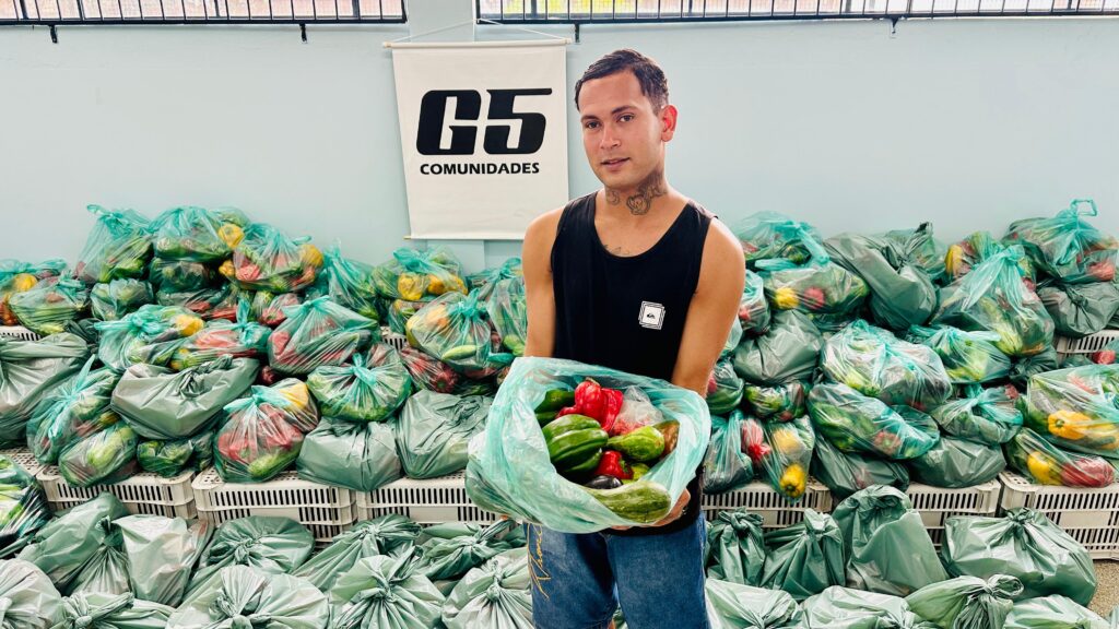 A imagem mostra um jovem homem em pé, segurando um grande saco plástico transparente cheio de legumes frescos, como pimentões vermelhos e verdes, e abobrinhas. Atrás dele, há uma enorme pilha de sacos plásticos semelhantes, todos contendo legumes, empilhados sobre caixas plásticas brancas. O homem está vestido com uma regata preta e jeans azuis, e tem tatuagens visíveis no pescoço e no peito. Ele está em um ambiente interno com paredes azuis claras e uma janela alta com grades. No fundo, há uma faixa ou banner com o texto "G5 COMUNIDADES". A cena sugere uma ação comunitária, possivelmente uma distribuição de alimentos para a comunidade.