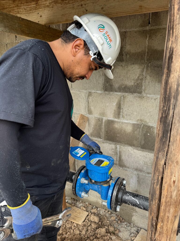 O homem, usando um capacete branco com o logotipo "SANE SOLUTI" em laranja e verde, está concentrado em seu trabalho. Ele usa uma camiseta preta com mangas compridas e luvas azuis com detalhes amarelos. Em suas mãos, ele segura uma chave inglesa, enquanto seus olhos estão fixos em um medidor de água azul, que está conectado a um tubo preto. O medidor possui uma tampa azul aberta, revelando uma tela digital e alguns rótulos amarelos. O fundo da imagem mostra uma parede de blocos de concreto e vigas de madeira. O chão está coberto de terra e alguns pedaços de madeira. Há uma quantidade visível de poeira ou detritos caindo sobre a camiseta do homem e o capacete.