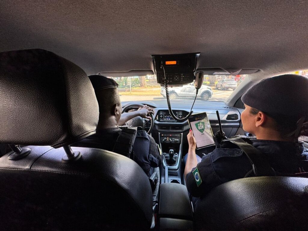 A imagem mostra dois policiais dentro de um carro, ambos usando uniformes escuros e boinas. O policial do lado do motorista está com as mãos no volante, enquanto a policial do lado do passageiro segura um tablet que exibe um aplicativo com um símbolo de SOS, e tem um rádio comunicador em sua mão. No teto do carro, há um rádio comunicador fixado, com um microfone pendurado. A cena sugere que eles estão em serviço, possivelmente em patrulha ou respondendo a uma ocorrência. A bandeira do Brasil está visível na manga do uniforme da policial.