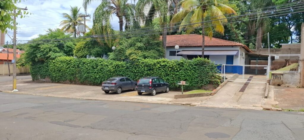 * A imagem mostra a fachada de um edifício com uma cerca viva alta e densa que o circunda. * Dois carros estão estacionados em frente à cerca viva: um cinza à esquerda e um preto à direita. * O edifício tem um telhado de telhas vermelhas e paredes claras com detalhes em azul. Há uma entrada com portão metálico branco e uma rampa de acesso. * Palmeiras e outras árvores frondosas criam um ambiente tropical e sombreado. * A rua em primeiro plano está asfaltada e apresenta algumas irregularidades. Um poste de eletricidade com fios elétricos atravessa a parte superior esquerda da imagem.