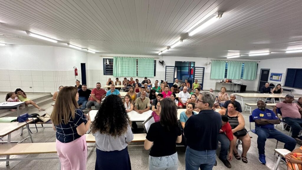 A imagem mostra uma sala com um grupo de pessoas sentadas em carteiras, como se estivessem em uma reunião ou aula. Há duas mulheres em primeiro plano, de costas para a câmera, que parecem estar falando com o grupo. Uma delas veste uma blusa quadriculada e calças cor de rosa, e a outra veste uma blusa listrada e calça escura. O grupo de pessoas sentadas é diversificado em idade e gênero, e algumas delas estão olhando para as mulheres que falam, enquanto outras parecem conversar entre si. Ao fundo, há janelas com cortinas verdes e algumas mesas com garrafas e outros objetos. O ambiente parece ser um local comunitário ou escolar, com paredes brancas e teto de lambris com iluminação fluorescente.