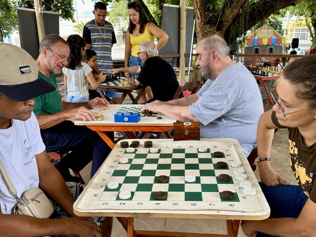 A imagem mostra um grupo de pessoas jogando xadrez e damas ao ar livre, sob a sombra de árvores. Há duas mesas com jogos em andamento. Em uma mesa, um homem mais velho com óculos joga xadrez com uma criança, enquanto um homem e uma mulher observam. Na outra mesa, um homem joga damas com outra pessoa, e um terceiro homem observa. As pessoas parecem estar relaxadas e aproveitando o momento. Ao fundo, há uma pequena estrutura que parece um palco de teatro de fantoches, com uma placa que diz "Teatro Fantoche". O ambiente é informal e parece ser um evento comunitário ou um encontro casual.