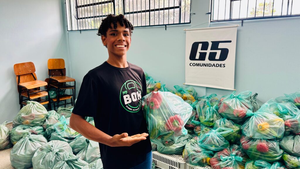 A foto mostra uma pessoa jovem, sorridente, com cabelo cacheado e escuro, vestindo uma camiseta preta com o logo "My BOX FITNESS • NUTRITION". Ela está segurando um saco plástico transparente cheio de pimentões vermelhos e verdes, e com a outra mão está fazendo um gesto aberto, como se estivesse apresentando ou explicando algo. Ao fundo, há várias sacolas plásticas verdes espalhadas no chão, também contendo pimentões, sugerindo uma ação de distribuição de alimentos. A parede atrás da pessoa é azul clara, com duas janelas gradeadas e uma faixa branca pendurada com o logo "G5 COMUNIDADES". Também é possível ver algumas cadeiras de madeira empilhadas. A imagem transmite uma atmosfera de trabalho comunitário, envolvendo alimentação saudável e apoio à comunidade.