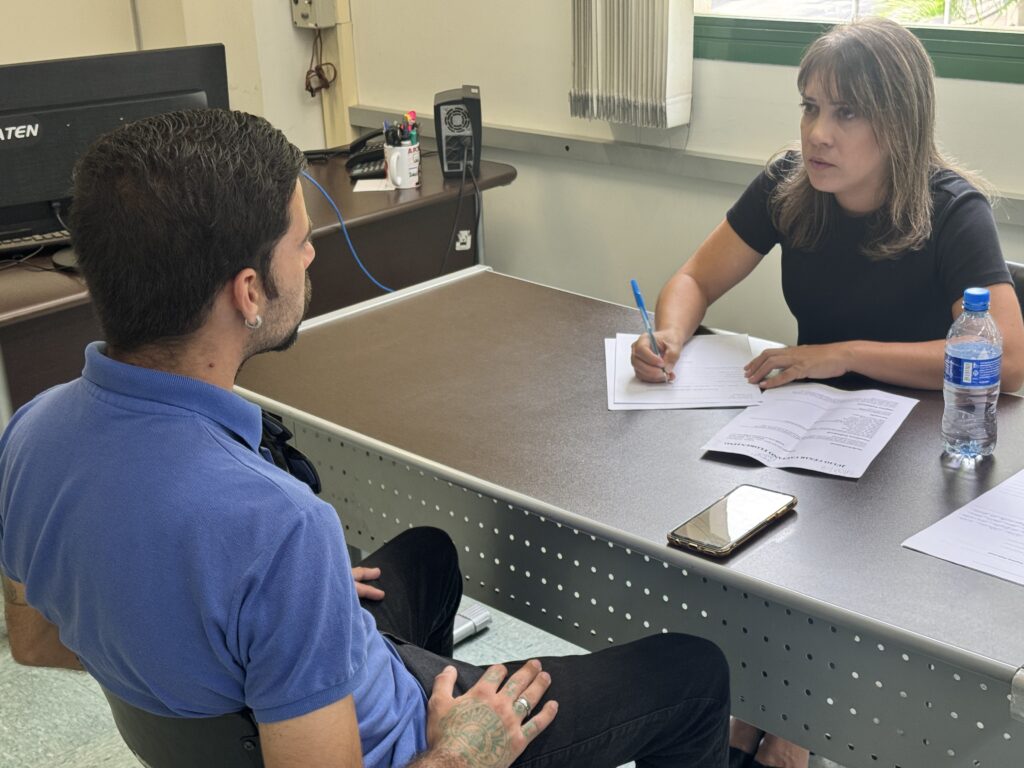 A imagem mostra duas pessoas sentadas em lados opostos de uma mesa de escritório. Uma mulher, com cabelo castanho e uma blusa preta, está escrevendo em um documento com uma caneta azul. Um homem, com cabelo escuro e uma polo azul, está sentado à sua frente, voltado para ela. Na mesa, há também uma garrafa de água, um celular e outros papéis. O ambiente parece ser um escritório, com um computador e outros objetos de escritório visíveis ao fundo.