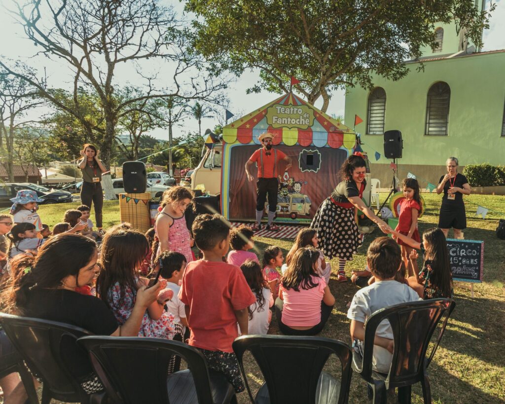 A imagem mostra uma cena ao ar livre com um grupo de pessoas, incluindo crianças e adultos, reunidas em torno de uma pequena tenda de teatro. A tenda é colorida e decorada com o nome "Teatro de Fantoche". Um palhaço com roupas vibrantes está em pé no palco da tenda, interagindo com a plateia. Uma mulher, também com maquiagem de palhaço, está do lado de fora da tenda, estendendo a mão para uma criança. Há também outros adultos e crianças sentados na grama, assistindo à apresentação. Ao fundo, vemos árvores, carros estacionados e um edifício verde. Uma placa escrita "CIRCO 15:00 HS GRÁTIS" sugere que o evento é uma apresentação de circo gratuita. A atmosfera geral é de diversão e entretenimento familiar.
