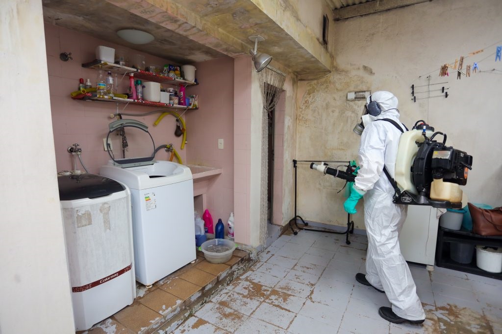 A imagem mostra uma pessoa vestindo um traje de proteção completo, incluindo um macacão branco, luvas verdes, máscara de gás e óculos de proteção, enquanto opera um pulverizador com mochila. A pessoa está em um cômodo que parece ser uma área de serviço ou lavanderia, com máquinas de lavar roupa, prateleiras com produtos de limpeza e uma pia. O chão está sujo e descascado em algumas áreas, e as paredes parecem desgastadas. Há também um varal improvisado com roupas penduradas no fundo. A pessoa está direcionando o bico do pulverizador para a parede, possivelmente realizando um processo de desinfecção ou dedetização.