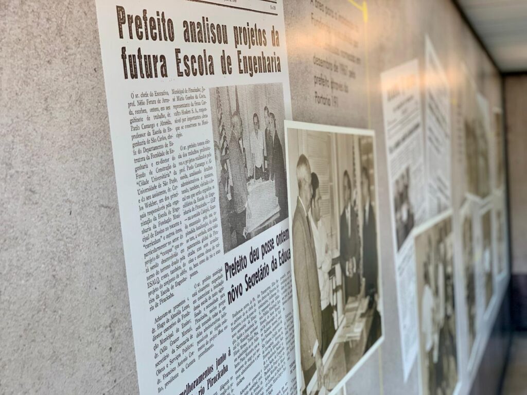 A imagem exibe uma parede com uma exposição de recortes de jornal e fotografias em preto e branco, dispostos em uma linha do tempo. O foco principal está em um jornal com a manchete "Prefeito analisou projetos da futura Escola de Engenharia", datado de 1968. O texto relata a visita do prefeito de Piracicaba, Nélio Ferraz de Arruda, acompanhado de representantes da firma Car-Talto Hosken S.A., para analisar os projetos da futura Escola de Engenharia. O artigo detalha a participação do professor Paulo Camargo e Almeida, chefe do Departamento de Estrutura da Faculdade de Engenharia e ex-diretor do Fundo de Construção da "Cidade Universitária da Universidade de São Paulo", e de seu assistente, Dr. Carlos Welcher, na organização da Escola de Engenharia da Fundação Municipal de Ensino. São mencionados os aspectos de "curriculum" e o projeto do "campus" que seria desenvolvido em torno do terreno doado pela ESALQ. Outro recorte de jornal, com a manchete "Prefeito deu posse ontem novo Secretário da Educação", também é visível, indicando a continuidade de eventos importantes na cidade. A fotografia ao lado mostra o prefeito e outras autoridades em um ambiente formal, possivelmente durante a posse do novo secretário. A exposição parece celebrar marcos históricos e o desenvolvimento da educação e infraestrutura em Piracicaba.