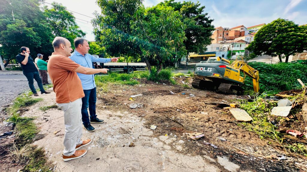 A imagem mostra um grupo de pessoas acompanhando uma obra ou ação de limpeza urbana, com destaque para uma escavadeira amarela em operação, removendo entulhos e vegetação. No primeiro plano, dois indivíduos apontam para a máquina, enquanto outros observam mais ao fundo, próximos a uma rua com veículos estacionados. O cenário inclui árvores e construções residenciais, indicando que a atividade ocorre em uma área habitada.
