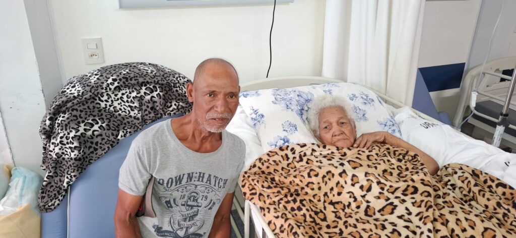 A imagem mostra dois indivíduos em um ambiente que parece ser um hospital ou clínica. À esquerda, um homem mais velho, com a pele bronzeada e cabelos grisalhos raspados, está sentado em uma cadeira ao lado de uma cama. Ele veste uma camiseta cinza com uma estampa que inclui um âncora e a frase "DOWN 'N HATCH". Ao lado dele, na cama, está uma mulher idosa, com cabelos brancos e cacheados, deitada e coberta com um cobertor de estampa de leopardo. Ela está apoiada em um travesseiro com estampa floral. O ambiente é iluminado e parece ser uma sala de internação, com cortinas brancas ao fundo e um suporte para soro ao lado da cama.