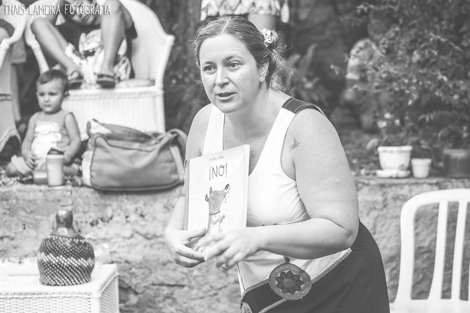 A imagem em preto e branco mostra uma mulher em um ambiente externo, segurando um livro intitulado "¡NO!" com uma ilustração de um cachorro na capa. Ela está em destaque, olhando levemente para o lado, com a boca aberta, como se estivesse falando ou contando algo. A mulher usa um top sem mangas e tem o cabelo preso com um grampo. Ela também usa um acessório redondo pendurado na cintura, que parece decorativo. Ao fundo, há uma criança sentada, olhando em direção à câmera, e alguns objetos, como uma cadeira de vime, plantas em vasos e bolsas. A foto transmite uma atmosfera de contação de histórias ou ensino, com foco na mulher e no livro que ela segura. O nome da fotógrafa, Thais Lameira, está visível no canto superior esquerdo da imagem.