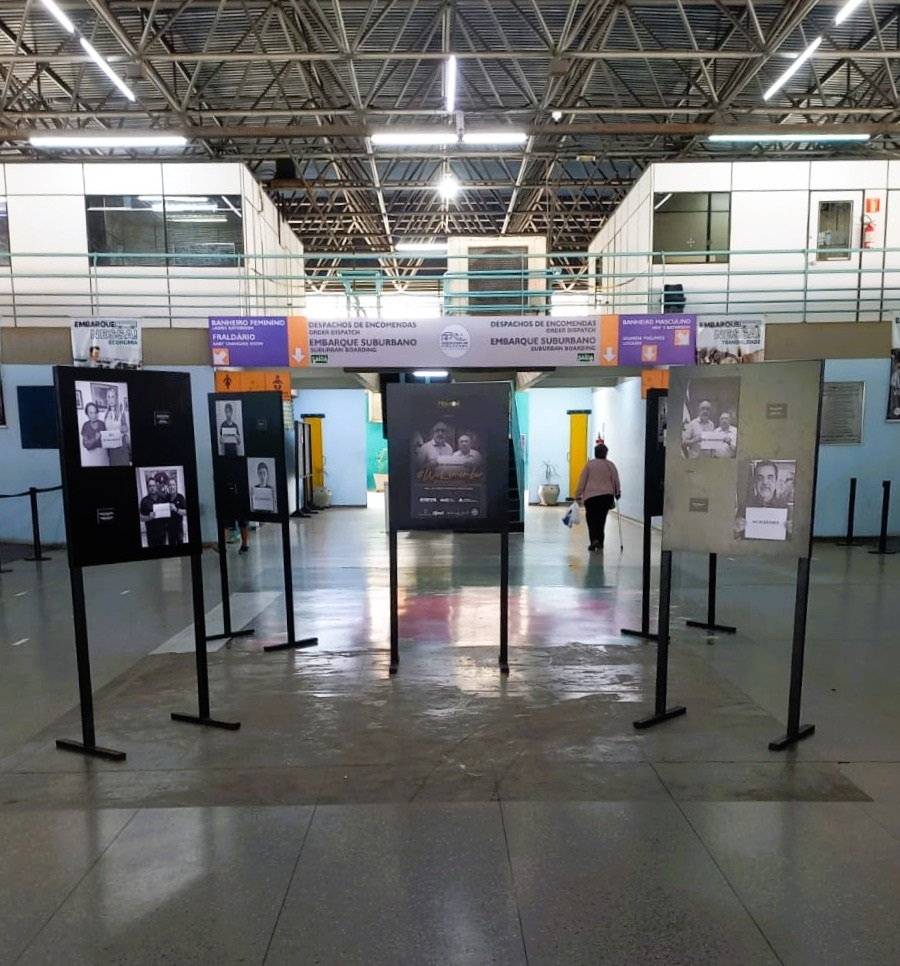 A imagem mostra uma exposição de fotografias em um espaço interno, possivelmente uma estação de trem ou um terminal. Há várias fotografias em preto e branco expostas em suportes pretos. Ao fundo, há sinalização indicando "EMBARQUE SUBURBANO", "DESPACHOS DE ENCOMENDAS", "BANHEIRO FEMININO" e "FRALDARIO". Uma pessoa caminha pela área, carregando sacolas e usando uma bengala. O ambiente é amplo, com teto alto e estrutura metálica aparente, iluminado por luzes artificiais.