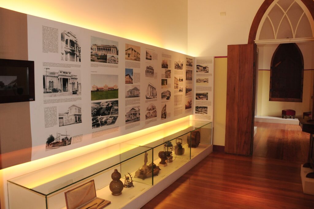 A imagem mostra uma exposição em um museu ou galeria. Em primeiro plano, há um longo display de vidro com várias peças de arte ou artefatos históricos expostos. Os objetos dentro do display parecem ser feitos de metal e têm um acabamento escuro. O display é iluminado por baixo por uma luz amarela quente e brilhante que realça os objetos. Atrás do display, há uma parede longa e branca que serve como tela para a exposição. A parede é coberta por uma série de fotografias e textos emoldurados, dispostos em uma grade. As fotografias parecem ser imagens históricas de edifícios, talvez documentando a arquitetura ou a história de uma determinada área. A iluminação na parte superior da parede também é amarela, criando uma atmosfera calorosa e convidativa. À direita da parede de exposição, há uma grande porta de madeira com um design antigo. Ao lado da porta, há uma abertura com uma janela arqueada em estilo gótico, que leva a outra sala. Essa sala adjacente tem piso de madeira escura e uma pequena área com assentos vermelhos, sugerindo que pode ser uma sala de estar ou uma área de descanso. O piso de madeira em toda a cena reflete a luz, adicionando ao brilho geral do espaço.