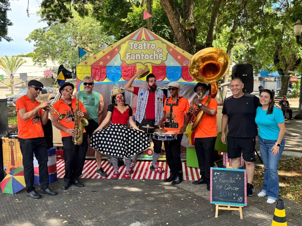 A imagem mostra um grupo de artistas de circo e músicos em frente a uma tenda com a inscrição "Teatro de Fantoche". Há dez pessoas visíveis na foto, a maioria vestida de forma colorida e festiva. No centro da imagem, uma artista com maquiagem de palhaço, usando um vestido preto com bolinhas brancas e meias listradas, está com a boca aberta em uma expressão animada. Ao lado dela, um homem com chapéu de palha e maquiagem de palhaço segura um chapéu em sua mão. À esquerda, três homens estão posicionados. Dois deles, vestidos com camisetas laranja e chapéus pretos, tocam instrumentos de sopro: um trompete e um saxofone. Um terceiro homem, com uma camiseta verde, observa a cena. À direita, outro homem vestindo uma camiseta laranja toca um tuba. Atrás dele, um homem e uma mulher, vestidos casualmente, sorriem para a câmera. Há também um alto-falante preto e uma pequena lousa com a inscrição "Kombi no Circo 10 H". O cenário é ao ar livre, com árvores frondosas ao fundo e uma rua de paralelepípedos no primeiro plano. A atmosfera geral é de alegria e celebração, sugerindo um evento de circo ou apresentação pública.