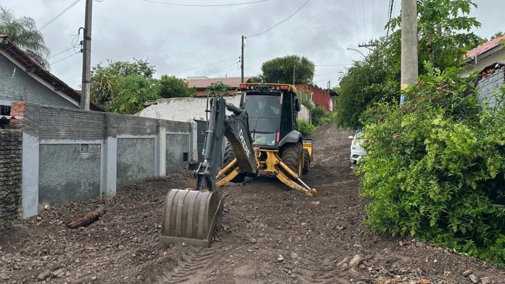 A imagem mostra uma retroescavadeira amarela e preta da marca John Deere posicionada em uma estrada estreita e enlameada. A máquina está com os estabilizadores abaixados, indicando que está em operação ou parada para algum trabalho. No primeiro plano, há uma grande concha metálica da escavadeira apoiada no chão. À esquerda, há um muro de concreto com painéis rebaixados, e à direita, vegetação densa e verde. Ao fundo, é possível ver árvores, um carro branco estacionado e postes de energia elétrica com fios. O céu parece nublado, criando uma iluminação difusa. A cena sugere que a retroescavadeira está sendo usada para manutenção ou nivelamento da estrada em uma área residencial ou semi-rural.