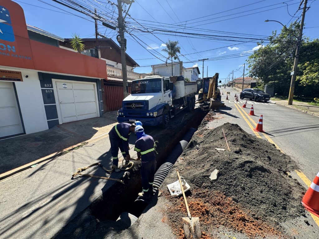 A imagem mostra uma cena de trabalho em uma rua residencial sob um céu azul claro. Há uma vala aberta no asfalto, onde dois trabalhadores vestindo uniformes azuis e capacetes brancos estão agachados, aparentemente instalando um tubo corrugado preto. Uma pá e uma enxada estão próximas à vala, e uma grande pilha de terra escura está ao lado. Um caminhão de despejo branco e azul, com a marca "CICAT", está estacionado ao lado da vala, com sua caçamba levantada. Atrás do caminhão, uma escavadeira amarela está posicionada, com seu braço mecânico sobre a vala. Na calçada à esquerda, um prédio de dois andares tem uma fachada laranja e branca com a marca azul de uma empresa. Um portão de garagem branco está visível, e placas com os números 823 e 819 estão fixadas na parede. A rua está marcada com linhas amarelas duplas e há cones de trânsito laranja espalhados, indicando a área de trabalho. Um carro escuro está estacionado mais adiante na rua, e postes de eletricidade com uma teia de fios cruzam o céu. Ao fundo, casas e árvores completam a paisagem urbana.