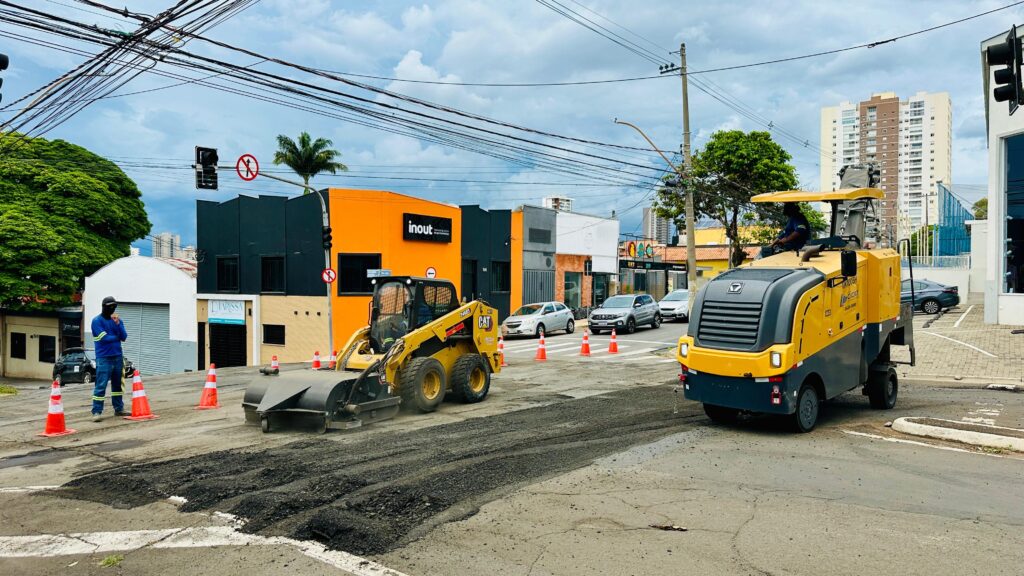 A imagem mostra uma cena de pavimentação de rua com dois veículos pesados em operação. À esquerda, um pequeno trator amarelo com uma lâmina de raspagem está sendo usado para remover o asfalto existente, com um trabalhador vestindo uniforme azul e máscara observando o trabalho. Vários cones de trânsito laranja estão dispostos ao redor da área de trabalho para sinalizar o perigo. À direita, um veículo amarelo e cinza, que parece ser um compactador de asfalto ou uma máquina de pavimentação, está em operação. Um operador está visível na cabine. O asfalto raspado está espalhado na rua, indicando que o trabalho está em andamento. Ao fundo, prédios comerciais com fachadas coloridas (laranja, preto e branco) estão alinhados na rua. Carros circulam nas faixas não afetadas pelo trabalho. Um prédio residencial alto se destaca na paisagem urbana ao longe. O céu está nublado, sugerindo um dia com possibilidade de chuva. A cena geral retrata um cenário urbano de manutenção de estradas.
