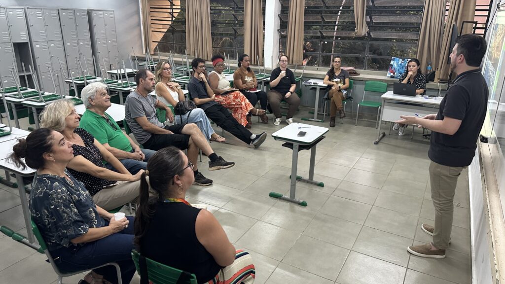 A imagem mostra um grupo de pessoas sentadas em uma sala de aula, aparentemente ouvindo uma apresentação. Um homem está de pé em frente a elas, com as mãos gesticulando, como se estivesse explicando algo. Ele está de costas para a câmera, mas parece estar apontando para um quadro ou tela que não está totalmente visível na imagem. As cadeiras e mesas da sala de aula estão empilhadas ao fundo, indicando que o espaço pode ser usado para diferentes propósitos. As pessoas sentadas parecem estar engajadas na apresentação, com expressões de atenção em seus rostos. A iluminação sugere que a sala está sendo iluminada pela luz natural que entra pelas janelas, além de iluminação artificial.