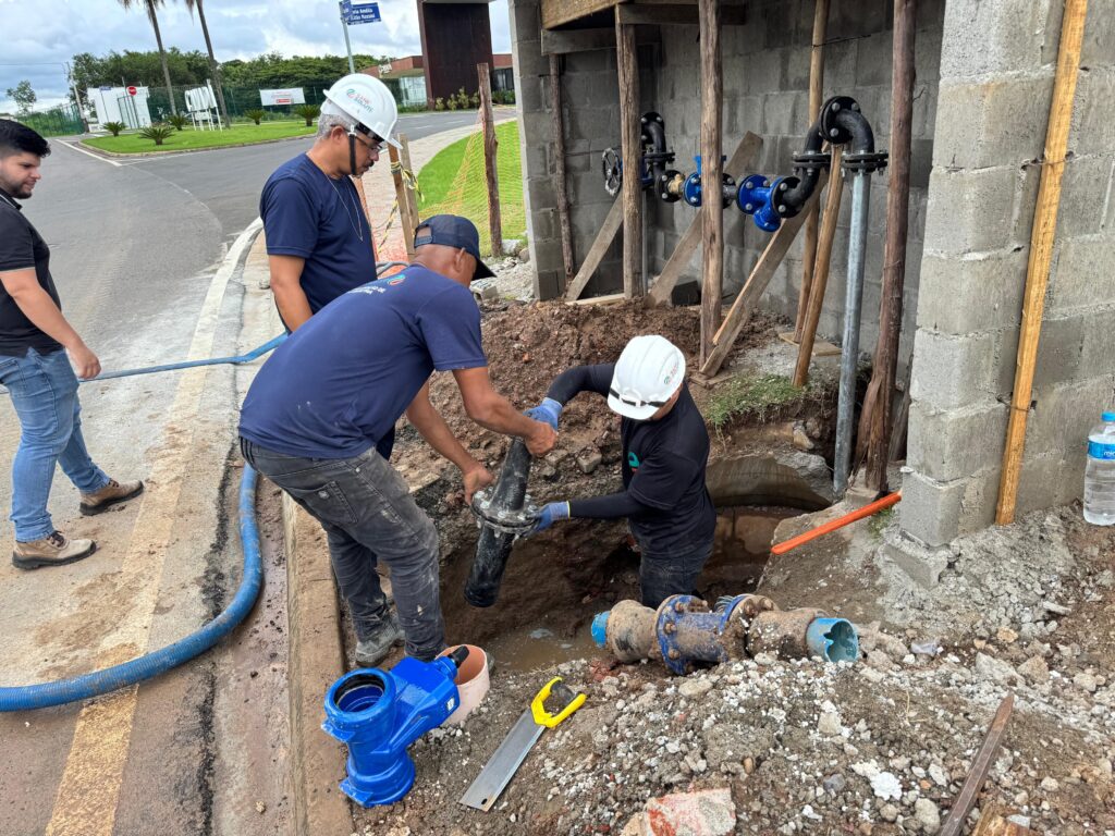 A imagem mostra uma equipe de trabalhadores realizando uma obra de encanamento. Quatro homens estão visíveis, três deles diretamente envolvidos na instalação ou manutenção de tubulações, enquanto um quarto observa ao fundo. No centro da imagem, dois trabalhadores estão agachados em uma vala, manuseando um grande cano preto com conexões metálicas. Um deles, usando um capacete branco e luvas azuis, segura a extremidade do cano enquanto o outro, vestindo um boné azul e luvas, auxilia na conexão. Ambos estão em meio a terra e entulho, indicando o local da obra. À esquerda, um homem de camiseta preta e jeans caminha próximo a uma mangueira azul grossa que se estende pela lateral da imagem. Mais atrás, um homem de capacete branco e camiseta azul escura observa a atividade, com uma corrente no pescoço. Ao fundo, pode-se ver uma estrutura de alvenaria com diversas conexões de tubulação aparentes, algumas delas pintadas de azul, sugerindo um sistema hidráulico complexo. A área ao redor é um canteiro de obras com terra, pedras e algumas ferramentas, como uma serra manual e um componente de encanamento azul brilhante. O céu nublado e a vegetação ao fundo indicam um ambiente externo.