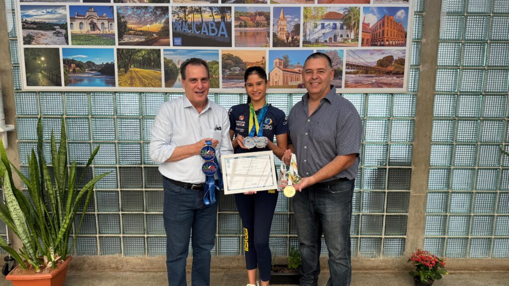A imagem mostra três pessoas em frente a uma parede decorada com fotos de paisagens. No centro, uma jovem atleta, vestindo um uniforme esportivo azul escuro com detalhes em amarelo, segura um certificado e exibe várias medalhas. Ela tem medalhas de prata presas ao pescoço e uma medalha de ouro pendurada em sua mão direita. Duas medalhas adicionais, com fitas azuis e inscrições "WORLD CUP", estão nas mãos do homem à esquerda. À esquerda da atleta, um homem mais velho, vestindo uma camisa social azul clara e calça jeans, segura duas medalhas com fitas azuis. Ele sorri para a câmera. À direita da atleta, um homem de meia-idade, com uma camisa azul escura com estampa de bolinhas brancas e calça jeans, também sorri. Ele segura uma medalha de ouro e outras duas medalhas. O fundo é composto por uma parede com um padrão de blocos de vidro translúcido e uma fileira de fotografias que retratam cenas da cidade de Piracicaba, incluindo edifícios históricos, pontes e paisagens naturais. Há também uma planta de espada-de-são-jorge em um vaso à esquerda e um pequeno vaso de flores vermelhas à direita. A iluminação geral sugere um ambiente interno bem iluminado.