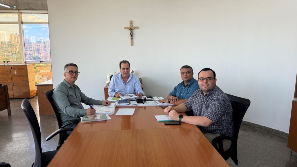 A imagem mostra quatro homens sentados em volta de uma mesa de madeira em uma sala de reuniões. Um homem está sentado à esquerda, usando óculos e uma camisa verde. Ele está escrevendo em um documento. O homem ao lado dele, no centro da imagem, está usando uma camisa azul clara e óculos. Ele está sorrindo e segurando alguns papéis. Atrás dele, um homem com uma camisa azul está sentado. O homem mais à direita está usando uma camisa xadrez e óculos, e também está sorrindo. Há uma cruz de madeira pendurada na parede atrás dos homens. A sala tem uma grande janela à esquerda, com vista para uma cidade. Há armários de madeira e uma planta na sala.