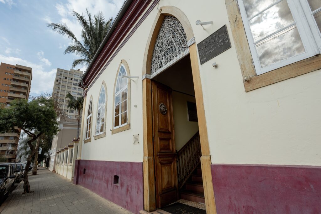 A imagem mostra a fachada de um edifício histórico, identificado por uma placa como o Museu H. P. Prudente de Moraes. O edifício possui uma arquitetura clássica, com paredes creme e uma base em tom vinho. Há três janelas em arco com molduras brancas e vitrais, e uma porta de madeira maciça com detalhes ornamentais. Acima da porta, uma grade elaborada complementa o design. À esquerda, uma calçada de pedras leva à entrada, ladeada por árvores e, ao fundo, prédios modernos e altos contrastam com a edificação histórica. O céu está parcialmente nublado, com a luz do sol refletindo em uma das janelas. A placa, em metal, contém a inscrição "MUSEU H. P. PRUDENTE DE MORAES NESTA CASA VIVEU E FALECEU O 1º PRESIDENTE CIVIL DA NAGAO".