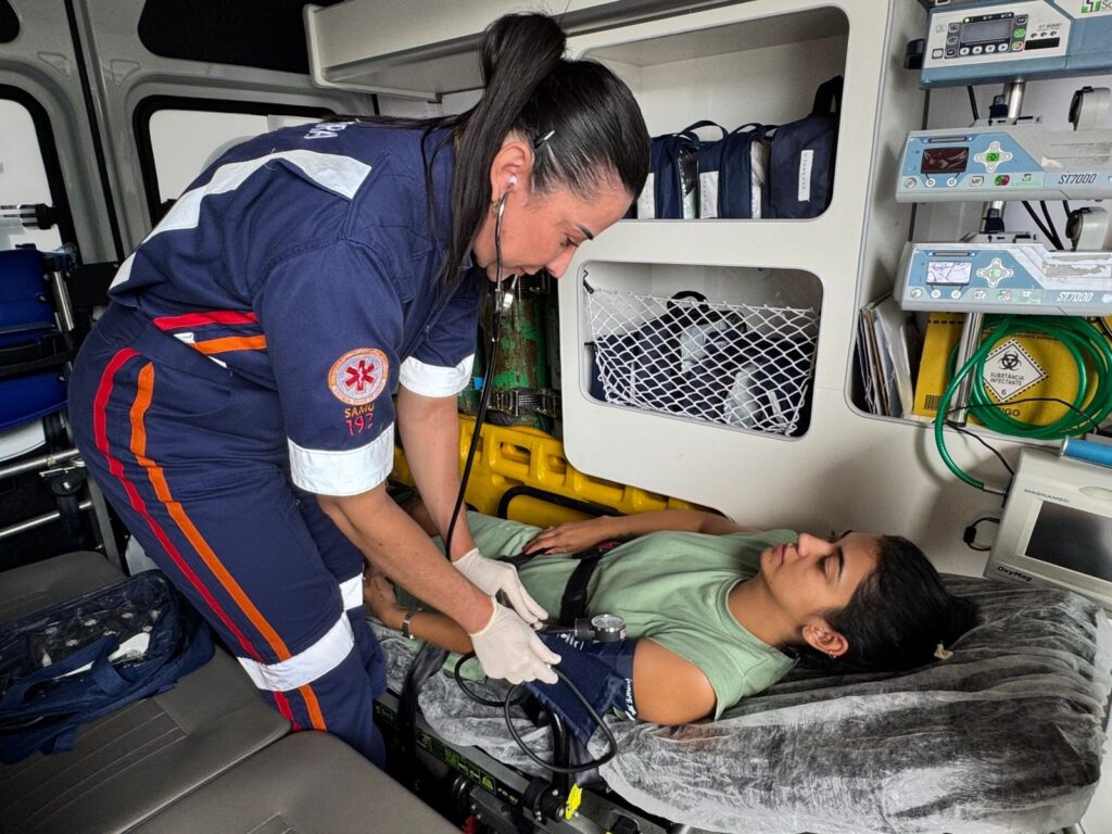 A imagem mostra uma profissional de saúde, vestida com um uniforme azul escuro com detalhes em laranja e branco, que parece ser do SAMU 192 (Serviço de Atendimento Móvel de Urgência). Ela está dentro de uma ambulância, agachada ao lado de uma maca. A profissional está examinando uma jovem paciente, que está deitada na maca. A profissional está usando luvas brancas e um estetoscópio, e está medindo a pressão arterial da paciente com um esfigmomanômetro. A paciente está vestida com uma camiseta verde e tem o cabelo escuro preso em um rabo de cavalo. O interior da ambulância está equipado com diversos aparelhos médicos, sacos de armazenamento e cilindros de oxigênio. A iluminação sugere que a foto foi tirada durante o dia.