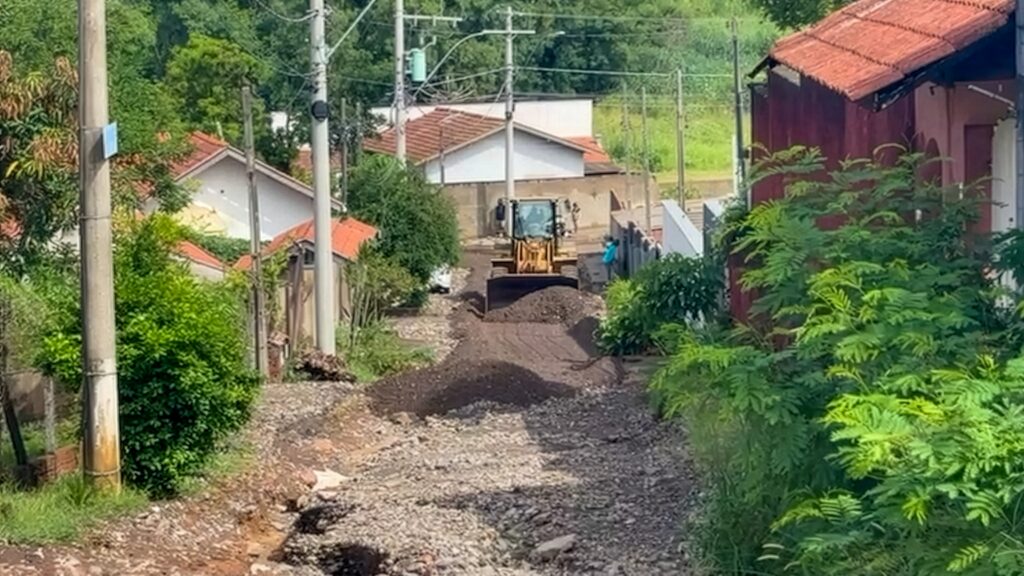 A imagem mostra uma cena de construção ou manutenção de uma estrada em uma área residencial arborizada. No centro da imagem, um trator amarelo está em operação, empurrando uma grande quantidade de terra escura. A estrada à frente do trator parece estar em processo de nivelamento ou alargamento, com pedras e terra espalhadas. Ao redor da estrada, há casas com telhados vermelhos e paredes claras, parcialmente obscurecidas pela vegetação exuberante. Postes de eletricidade com fios cruzam a cena, indicando a presença de infraestrutura urbana. Folhagens verdes e densas, incluindo árvores e arbustos, dominam as laterais da imagem, criando um contraste entre a obra em andamento e o ambiente natural. Algumas pessoas podem ser vistas perto do trator, possivelmente supervisionando ou participando do trabalho.