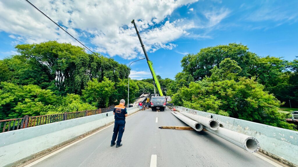 A imagem mostra uma ponte com uma equipe trabalhando em uma operação de remoção ou reparo. No centro da ponte, um grande guindaste verde está posicionado, com seu braço estendido em direção a uma estrutura danificada ou a ser removida. Uma caminhonete preta está posicionada sob o guindaste. Na frente do guindaste, há vários trabalhadores, um deles usando um colete laranja e preto com a inscrição "Proteção Defesa Civil". Ao lado da estrada na ponte, há três grandes tubos de concreto empilhados, apoiados em calços de madeira. O fundo da imagem é preenchido por uma densa vegetação verde e um céu azul com nuvens brancas. A atmosfera geral sugere uma operação de emergência ou de manutenção em andamento.