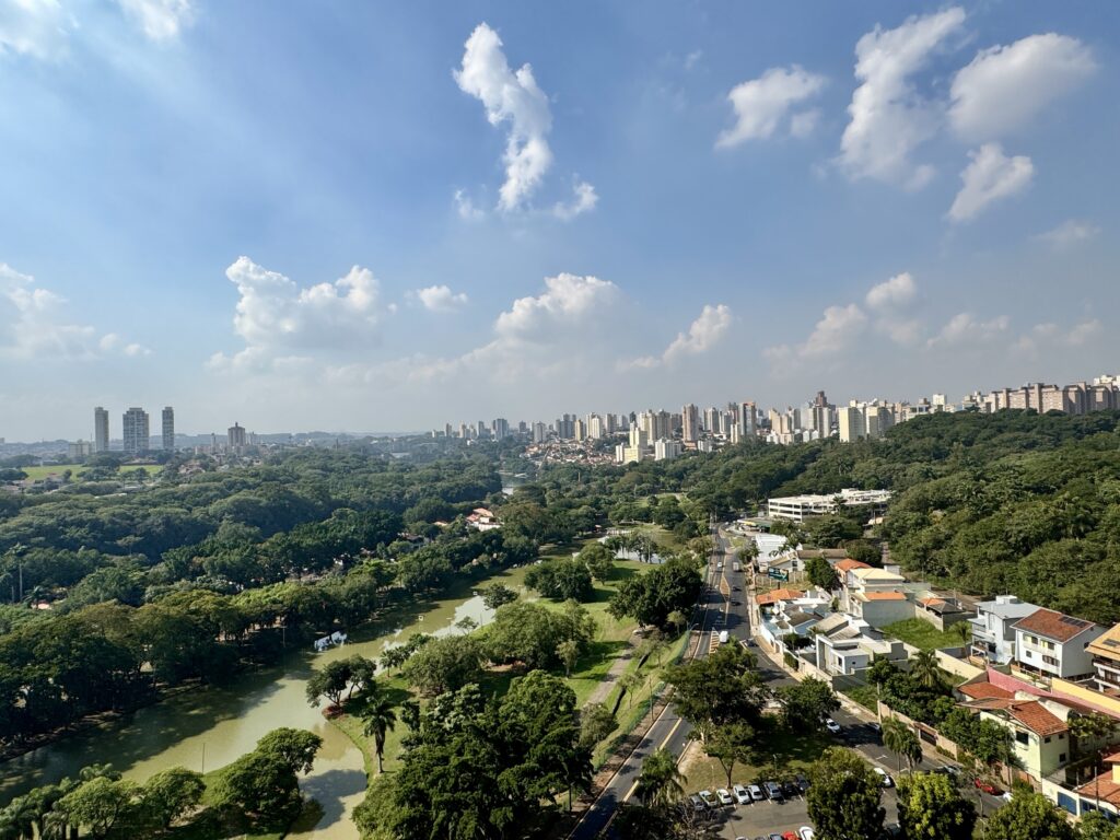 A imagem é uma paisagem urbana com muita vegetação, vista de um ponto elevado. No primeiro plano, à esquerda, vemos um lago de águas calmas e esverdeadas, cercado por árvores frondosas e gramados. Uma estrada sinuosa atravessa a paisagem, com casas de telhados vermelhos e brancos dispostas em um dos lados, e mais vegetação do outro. Ao fundo, estende-se uma vasta área verde que se funde com a cidade. Edifícios altos e modernos, com fachadas em tons claros, pontuam o horizonte, contrastando com as construções mais baixas e residenciais próximas à estrada. O céu azul, salpicado por nuvens brancas e fofas, completa a cena, conferindo uma atmosfera de tranquilidade e harmonia entre a natureza e o desenvolvimento urbano.
