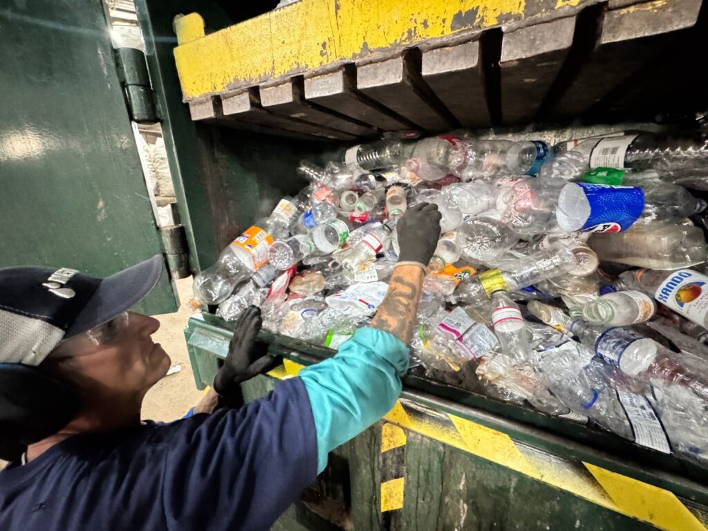 A imagem mostra um trabalhador em uma instalação de reciclagem, usando luvas pretas e um boné azul, empurrando garrafas plásticas para dentro de um compactador. O compactador é grande, com uma parte superior amarela e uma base verde. As garrafas plásticas estão amontoadas, com algumas marcas e rótulos visíveis, incluindo uma garrafa com um rótulo laranja e outra com um rótulo azul e branco. O trabalhador está usando um protetor auricular e óculos de segurança, indicando que ele está em um ambiente de trabalho barulhento e potencialmente perigoso. A cena sugere o processo de reciclagem de plástico, onde os materiais são coletados e compactados para processamento posterior.