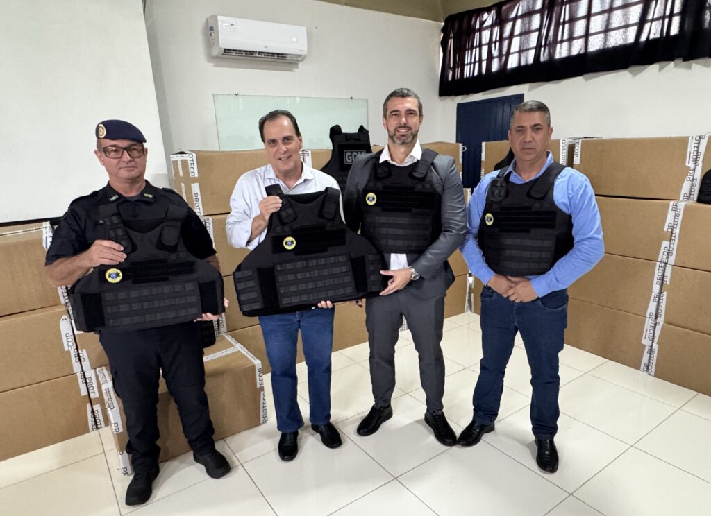 A imagem mostra quatro homens em um ambiente interno, provavelmente um escritório ou depósito, posando para a foto enquanto seguram ou usam coletes táticos balísticos. Eles estão alinhados lado a lado, de frente para a câmera. Ao fundo, há várias caixas de papelão empilhadas, todas lacradas com fita adesiva branca que tem a palavra "PROTECOP" impressa repetidamente. Os homens vestem roupas variadas: um está com uniforme tático preto e boina, outro usa camisa branca e jeans, o terceiro está de terno cinza com o colete balístico por cima, e o último usa camisa azul e jeans, também com o colete. No canto superior direito, há um aparelho de ar condicionado fixado na parede branca. A cena parece representar a entrega ou apresentação oficial dos coletes balísticos, possivelmente para uma equipe de segurança ou força policial, sugerido pela presença do homem com uniforme e a palavra "GCM" (Guarda Civil Municipal) visível nas caixas. A iluminação é clara e uniforme, destacando bem os detalhes dos coletes e das caixas ao fundo.