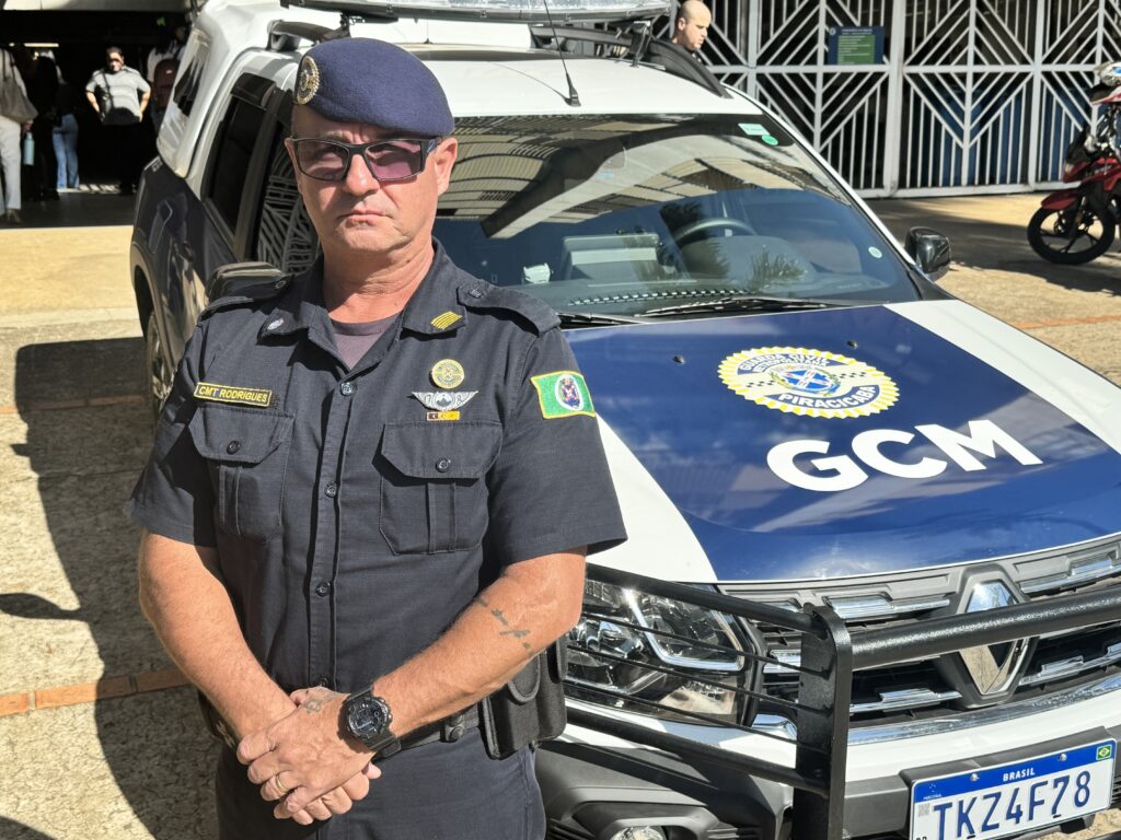 O homem, que parece ser um oficial da Guarda Civil Metropolitana (GCM), está em pé em frente a um carro de patrulha da GCM. Ele veste um uniforme azul escuro, incluindo um chapéu boina azul, óculos escuros e um distintivo no peito. O carro de patrulha é branco e azul, com o logotipo "GCM" na frente. Há outras pessoas e uma motocicleta em segundo plano, sugerindo que a cena se passa em um local público.