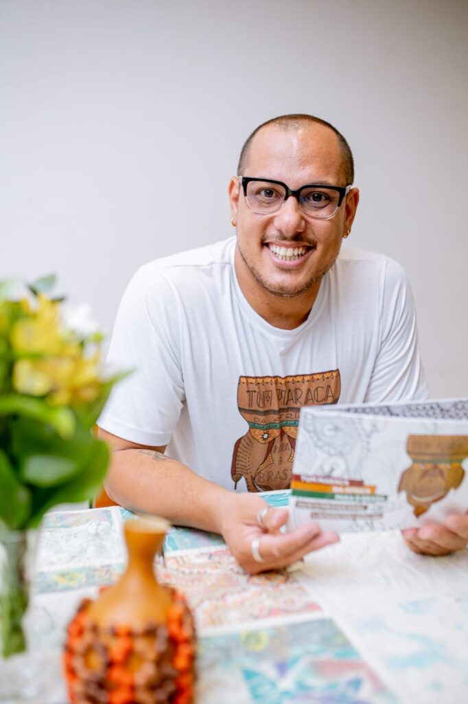 Um homem sorridente com óculos pretos está sentado à mesa, segurando um livro ou folheto. Ele usa uma camiseta branca com uma estampa gráfica que parece ser um desenho estilizado de uma figura com elementos culturais. A mesa tem uma toalha colorida e há um vaso com flores amarelas e brancas à esquerda, e um pequeno objeto decorativo marrom na frente. O homem tem anéis nos dedos e um brinco dourado na orelha. A iluminação é suave, destacando o rosto e as mãos do homem.