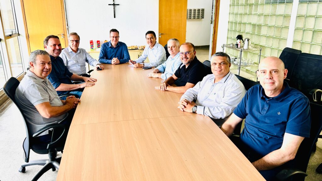 A imagem mostra um grupo de nove homens sentados ao redor de uma mesa de madeira em uma sala de reuniões. Eles parecem estar em um ambiente de escritório, com janelas grandes e uma divisória de vidro texturizado ao fundo. No lado esquerdo da mesa, vemos quatro homens. O mais próximo da câmera, à esquerda, usa uma camisa cinza e está sentado em uma cadeira preta giratória. Ao lado dele, um homem com camisa azul escura sorri para a câmera. Mais adiante, um homem com camisa clara e óculos está sentado, e um quarto homem com camisa azul está mais afastado. No lado direito da mesa, há cinco homens. O mais próximo da câmera, à direita, usa uma camisa azul marinho e está sentado em uma cadeira preta, olhando diretamente para a câmera. Ao lado dele, um homem com camisa clara e um relógio no pulso está com as mãos cruzadas. Mais adiante, um homem com camisa preta sorri. Os outros dois homens à mesa estão com camisas claras, um deles com óculos.